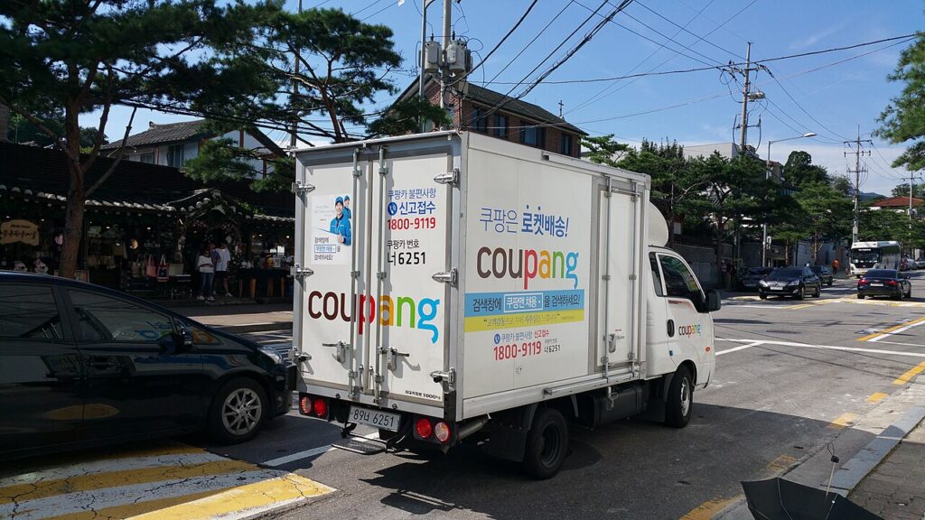 쿠팡 로고와 ‘로켓배송’ 문구가 크게 표시된 흰색 배송 트럭이 도로 위에 있는 모습